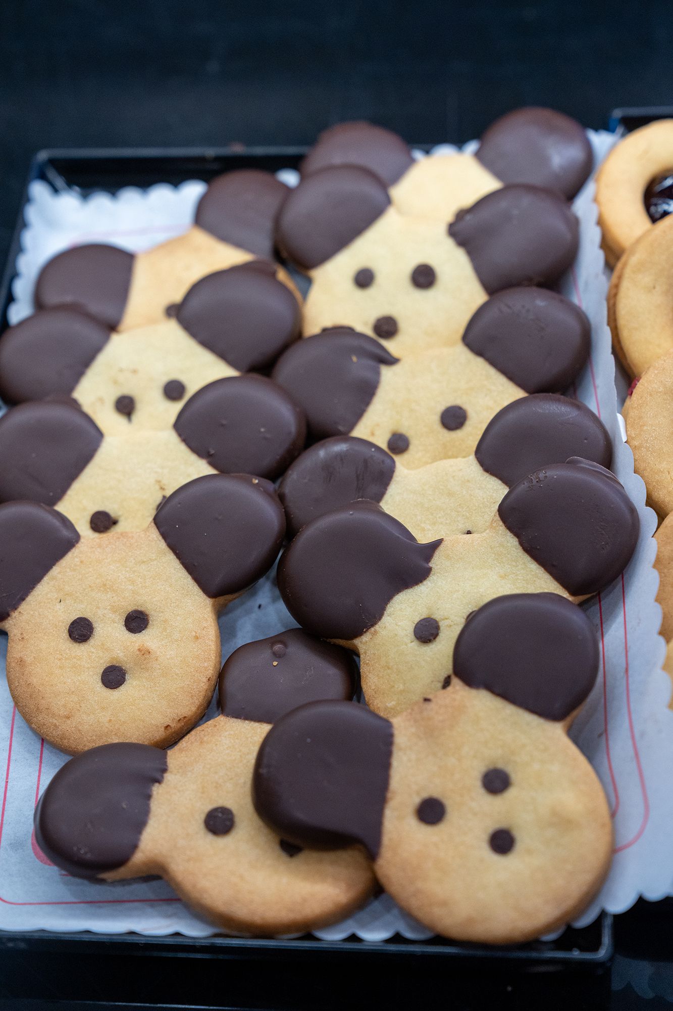 Vassoio di biscotti a forma di orsetto con orecchie ricoperte di cioccolato e due piccoli occhi di cioccolato su un centrino bianco.