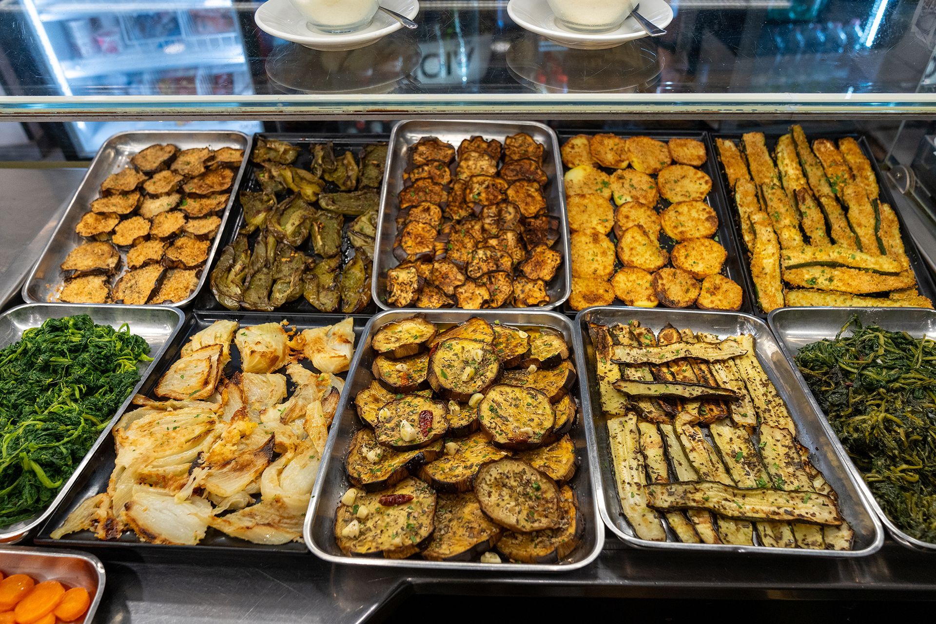 Esposizione di varie verdure cotte in vassoi di metallo su un banco alimentare.