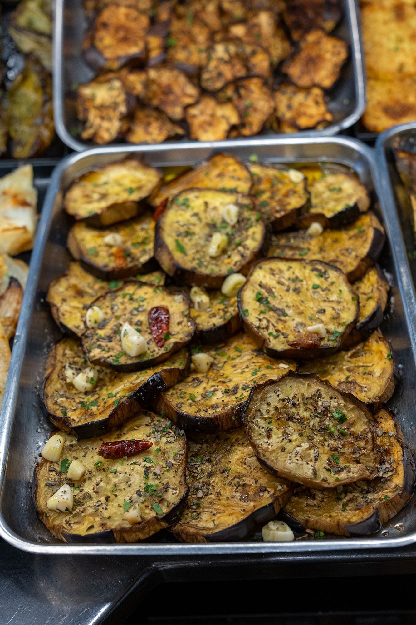 Fette di melanzane arrostite in una teglia di metallo, condite con erbe aromatiche, aglio e peperoncino.