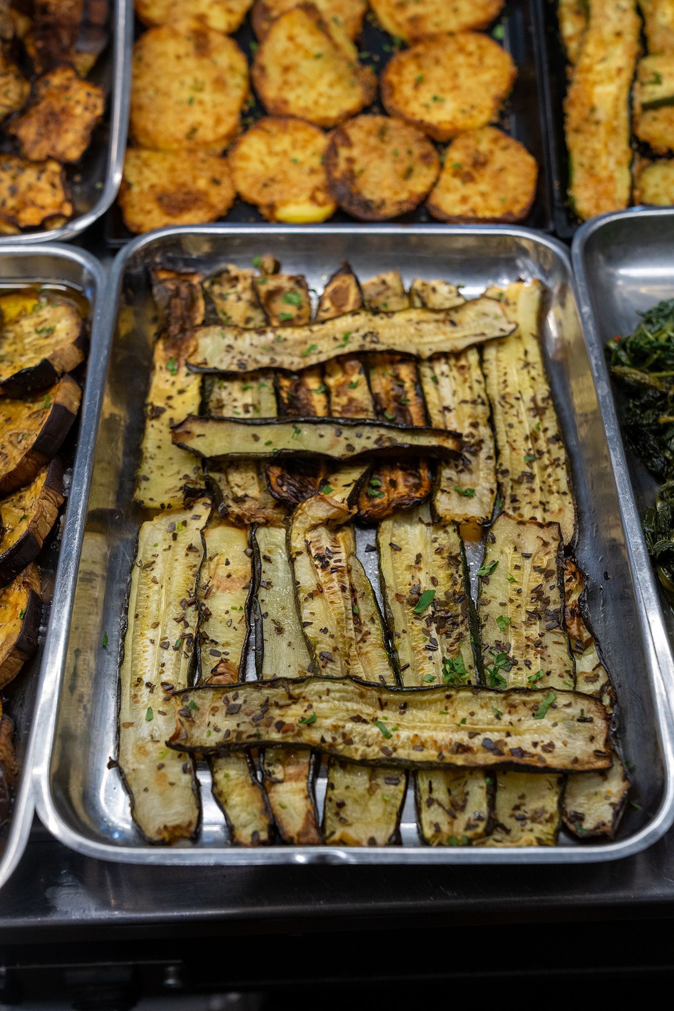 Fette di melanzane grigliate in un vassoio di metallo, esposte insieme ad altre verdure cotte al bancone del cibo.