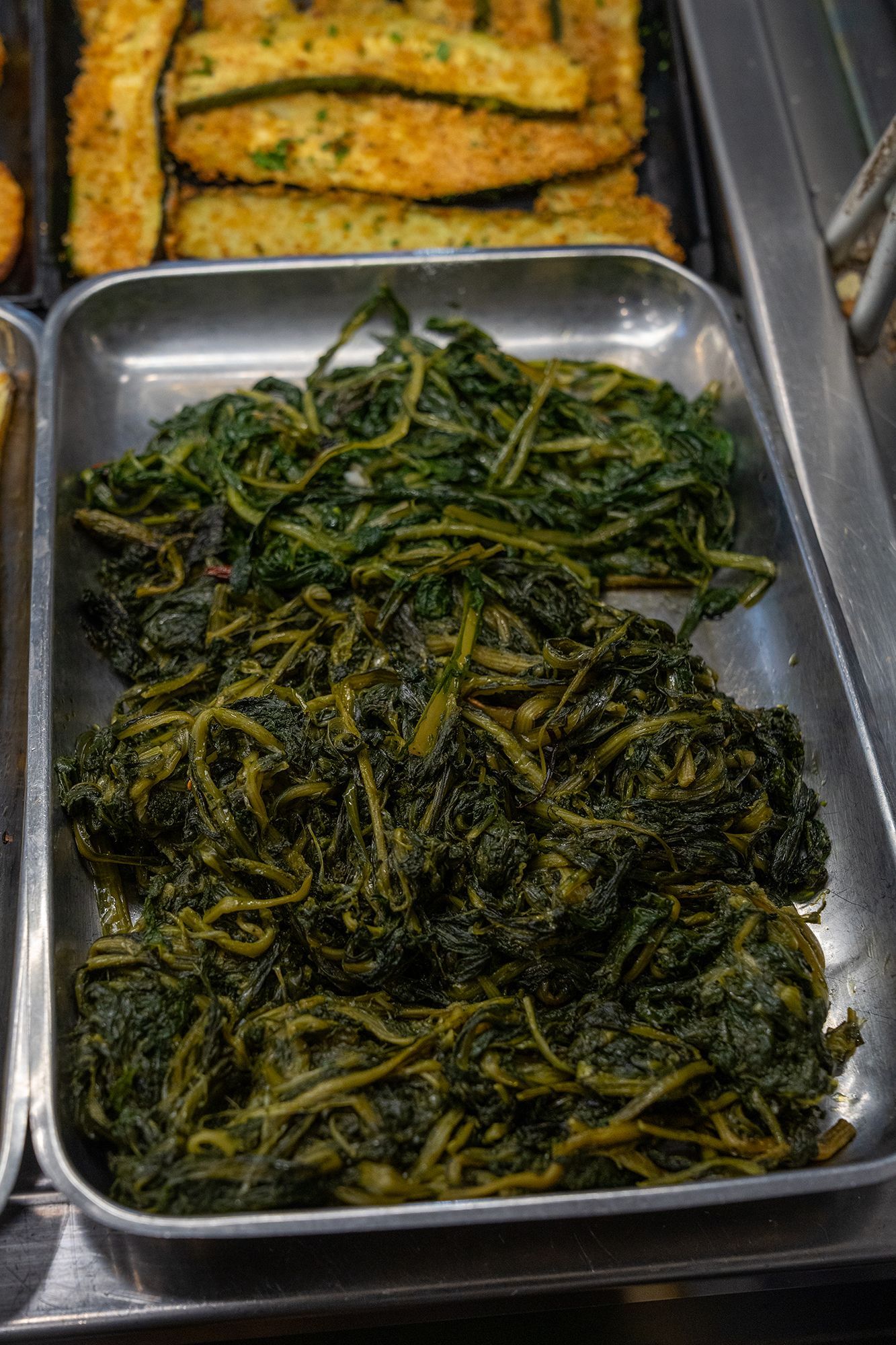 Due vassoi di verdure verdi cotte, possibilmente verdi o spinaci, esposti in un espositore per alimenti.