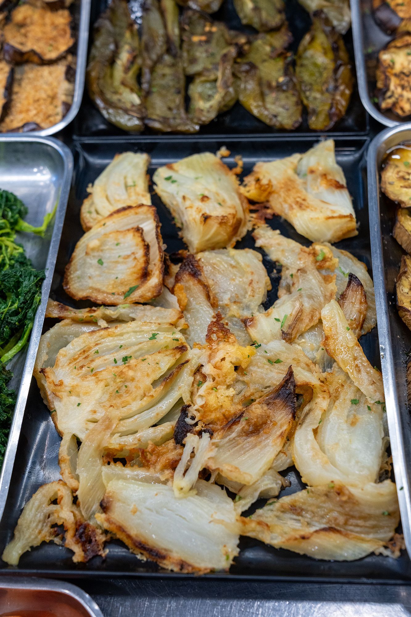Finocchio arrostito su un vassoio nero, con altre verdure cotte nei vassoi adiacenti.