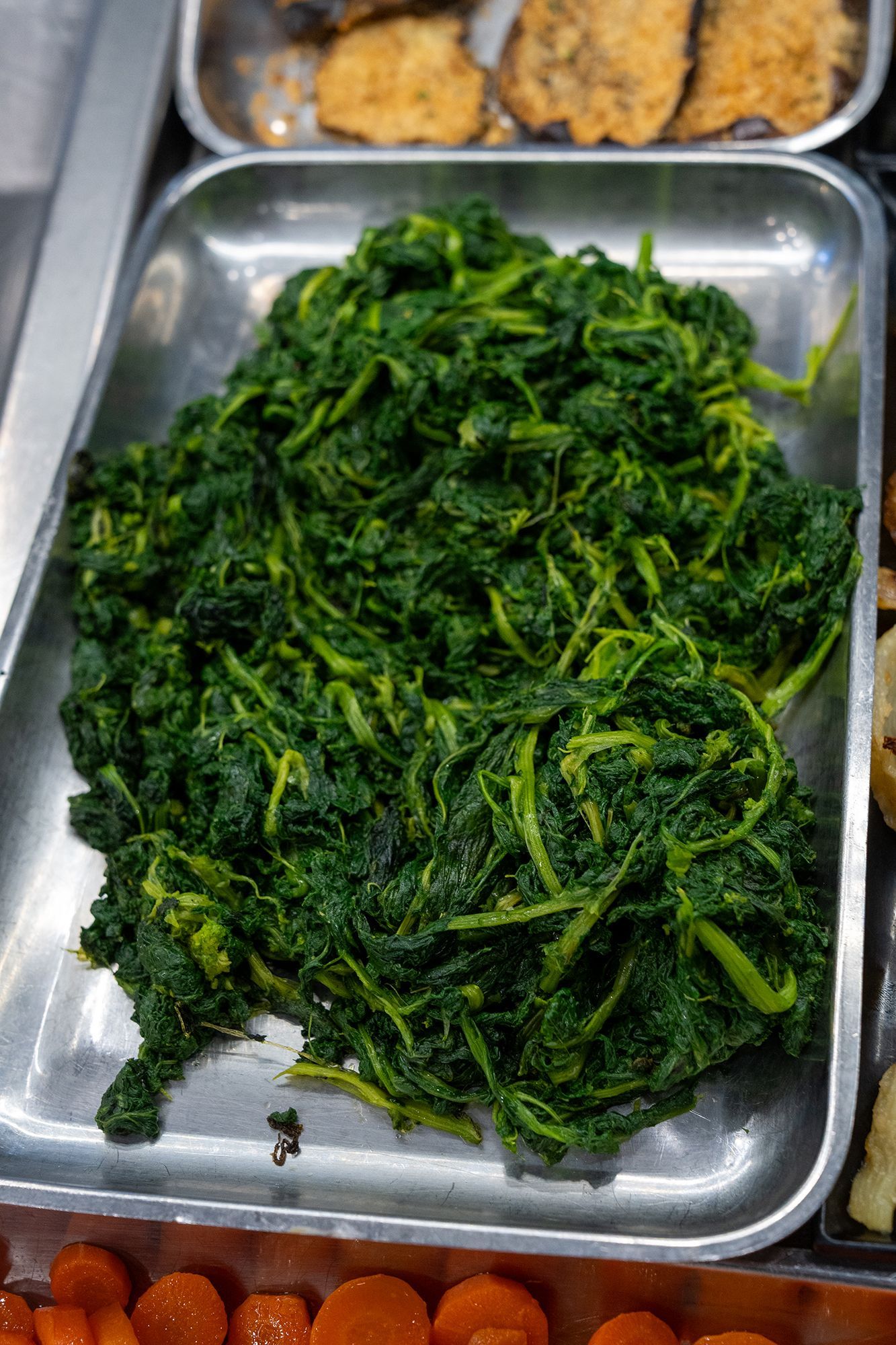 Mucchio di verdure verdi cotte in un vassoio di metallo, parte di un'esposizione di cibo.