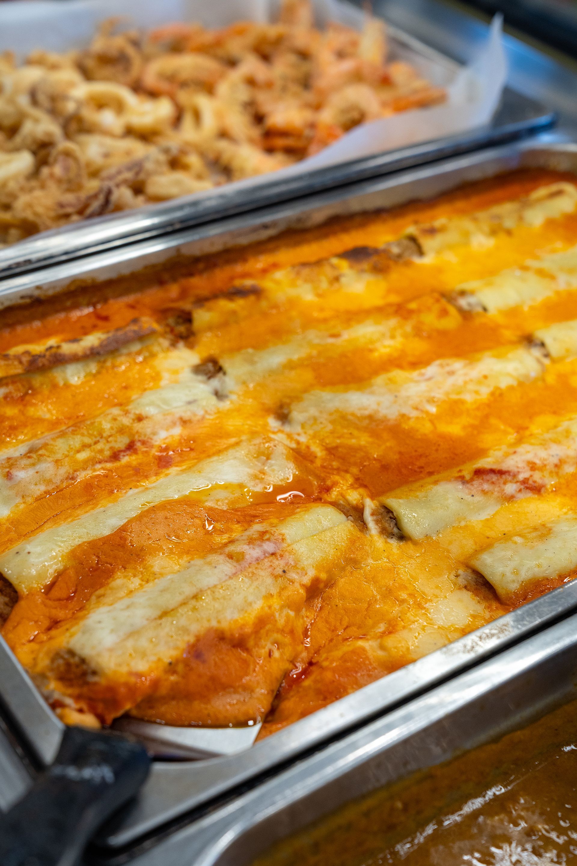 Teglia ripiena di involtini di pasta in salsa all'arancia, accanto a cibo fritto in un buffet.