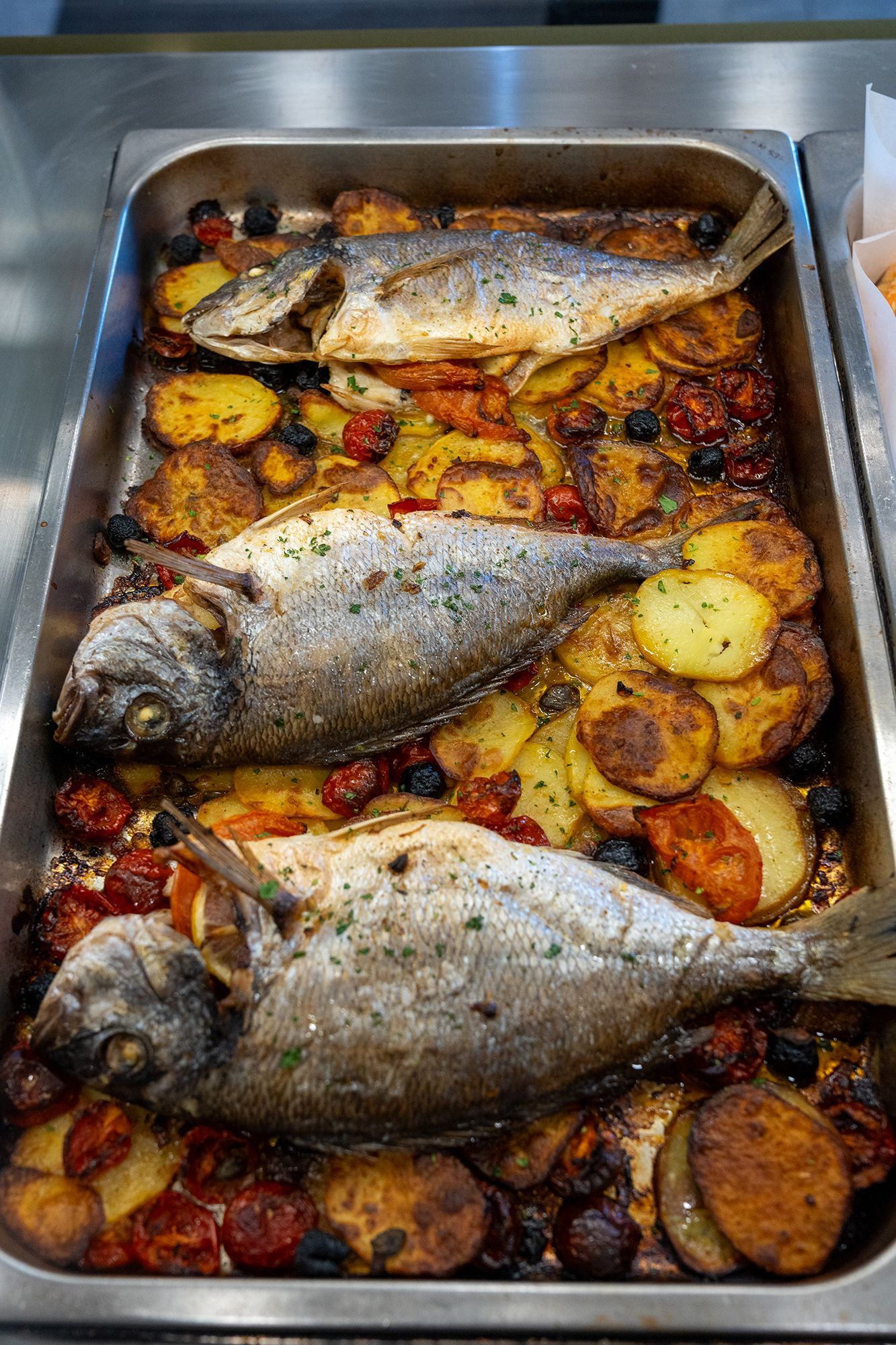 Pesce intero al forno con patate, pomodori e olive in una teglia di metallo.