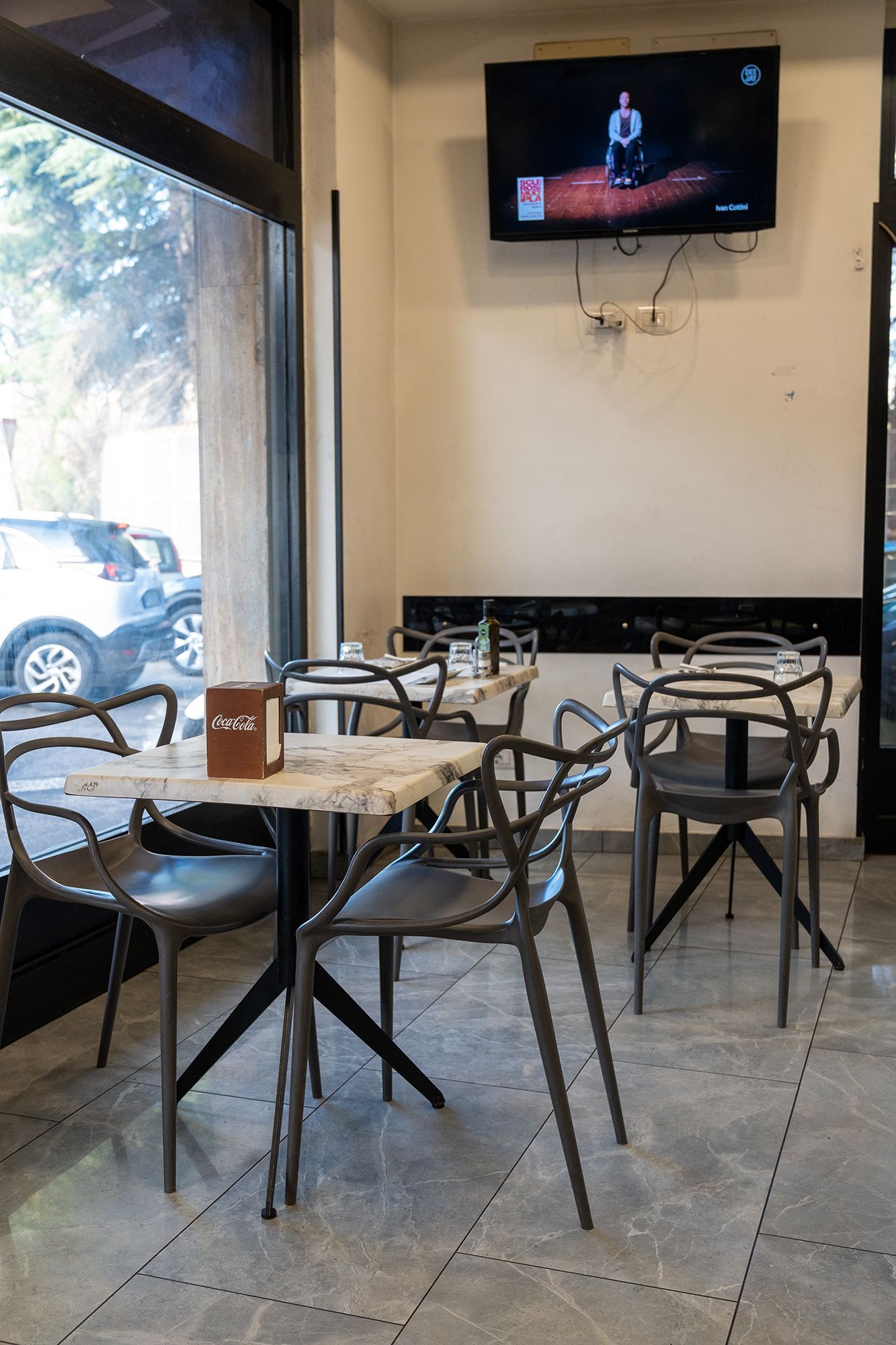 Bar interno con tavoli in marmo, sedie nere e una TV a parete.