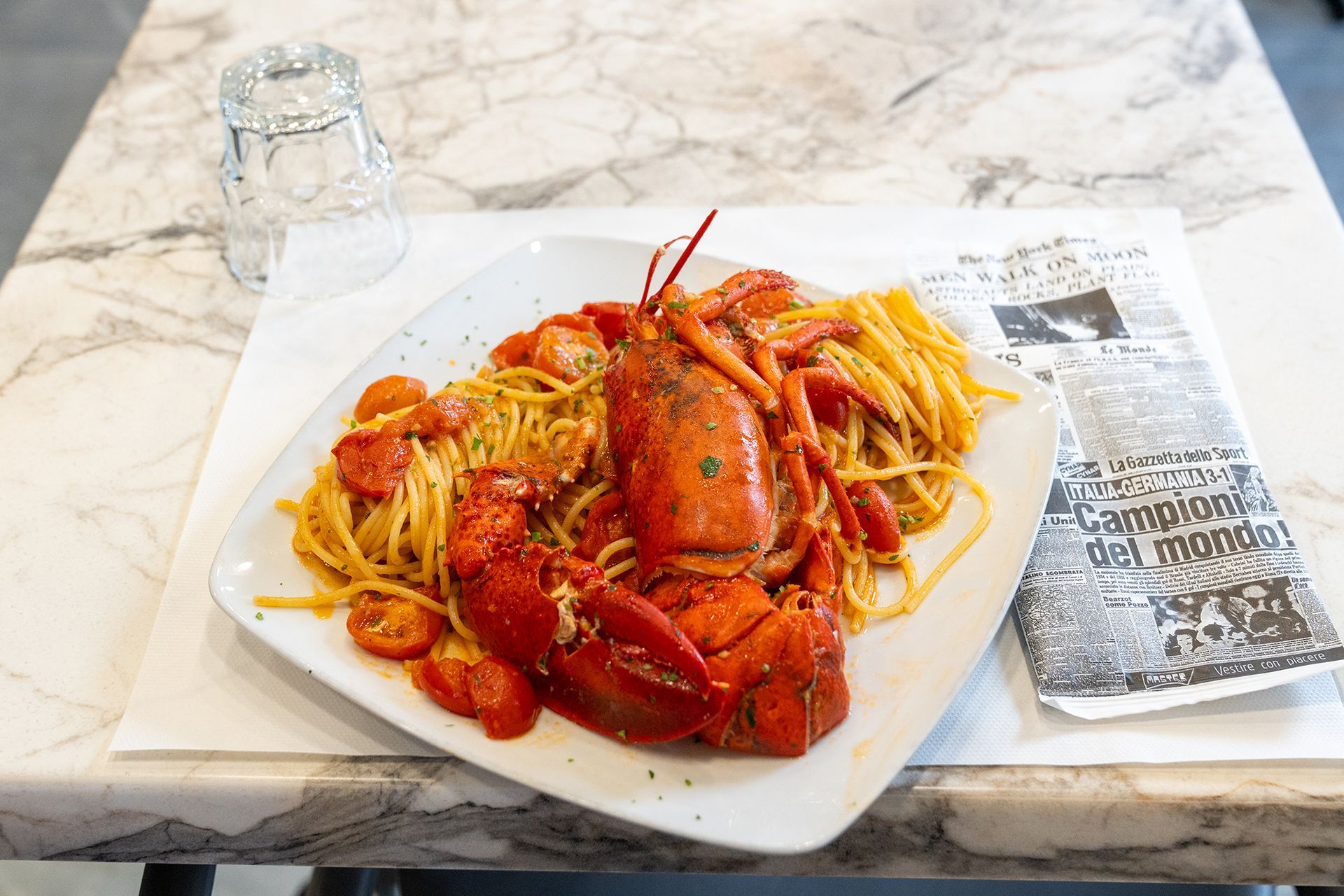 Piatto di pasta all'astice su un piatto bianco con un bicchiere d'acqua e un giornale su un tavolo di marmo.