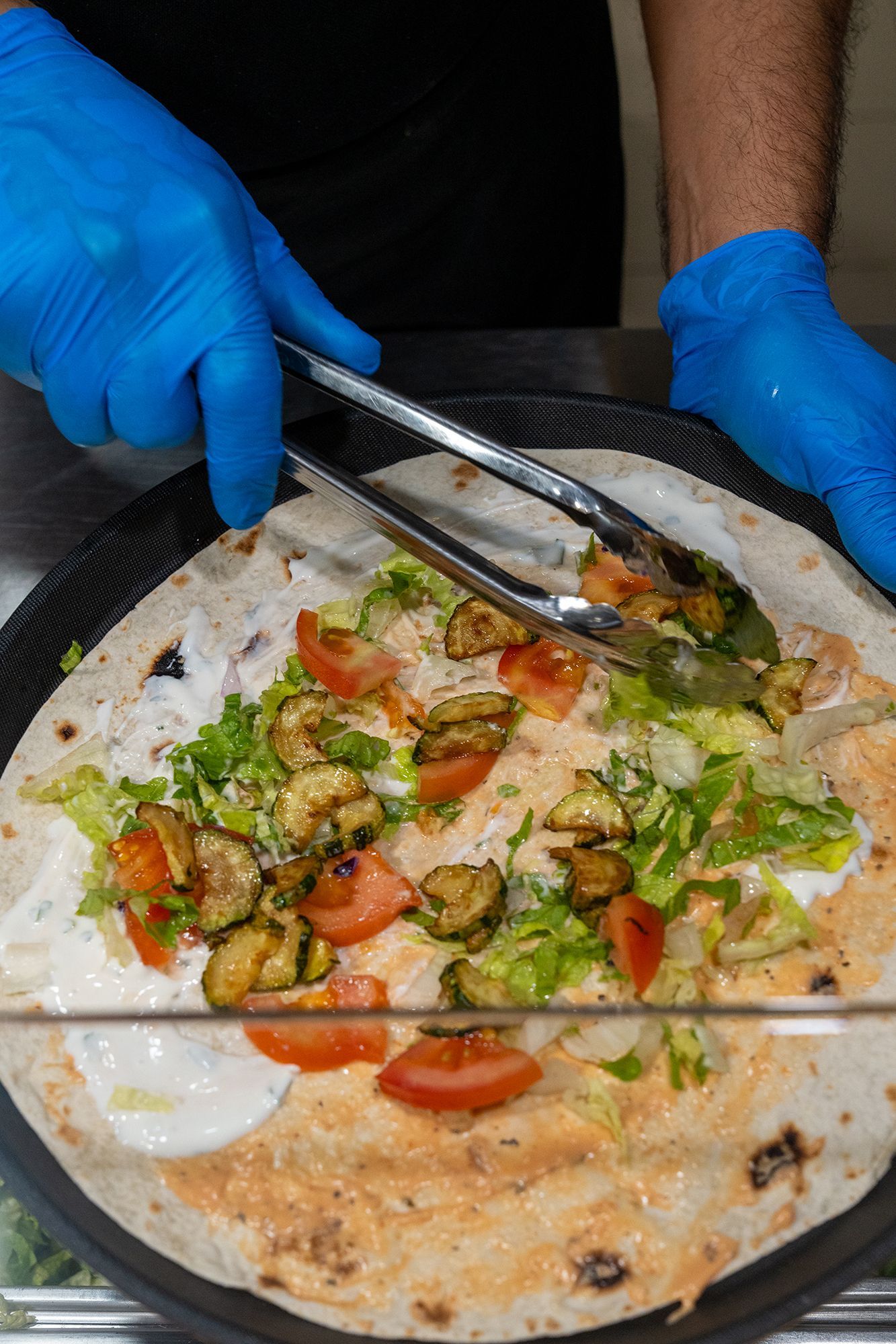 Una persona che indossa guanti blu e usa le pinze per comporre un wrap con verdure su una piadina.