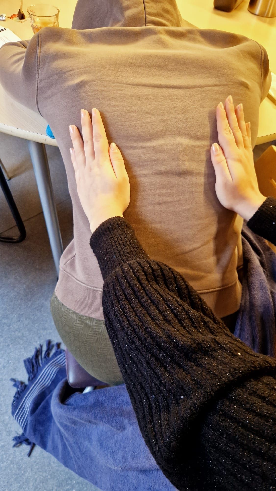 Handen masseren de rug van een persoon, gekleed in een bruine hoodie. De persoon zit binnen op een stoel.