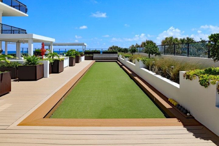 A bocce ball court with green turf, framed by wooden decking and white planters, set on an outdoor rooftop terrace.