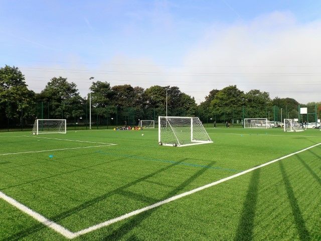 An outdoor synthetic soccer field with multiple white goal frames under a bright blue sky with scattered clouds.