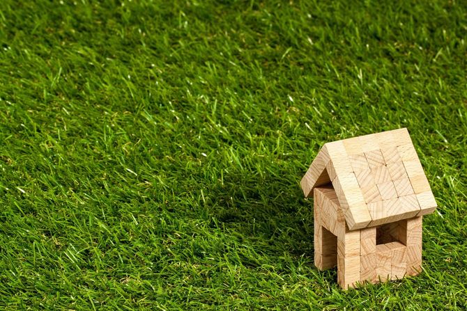 A small model house made of light-colored wooden blocks sits in the lower right corner on a patch of bright green grass.