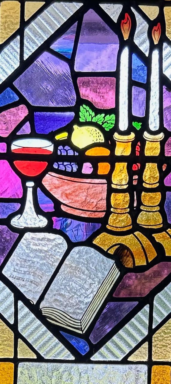 Stained glass window: chalice, fruit bowl, lit candles, and open book on a table.