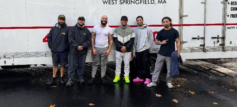 Six people pose outside a truck trailer marked “WEST SPRINGFIELD, MA,” wearing sports gear and neon shoes.