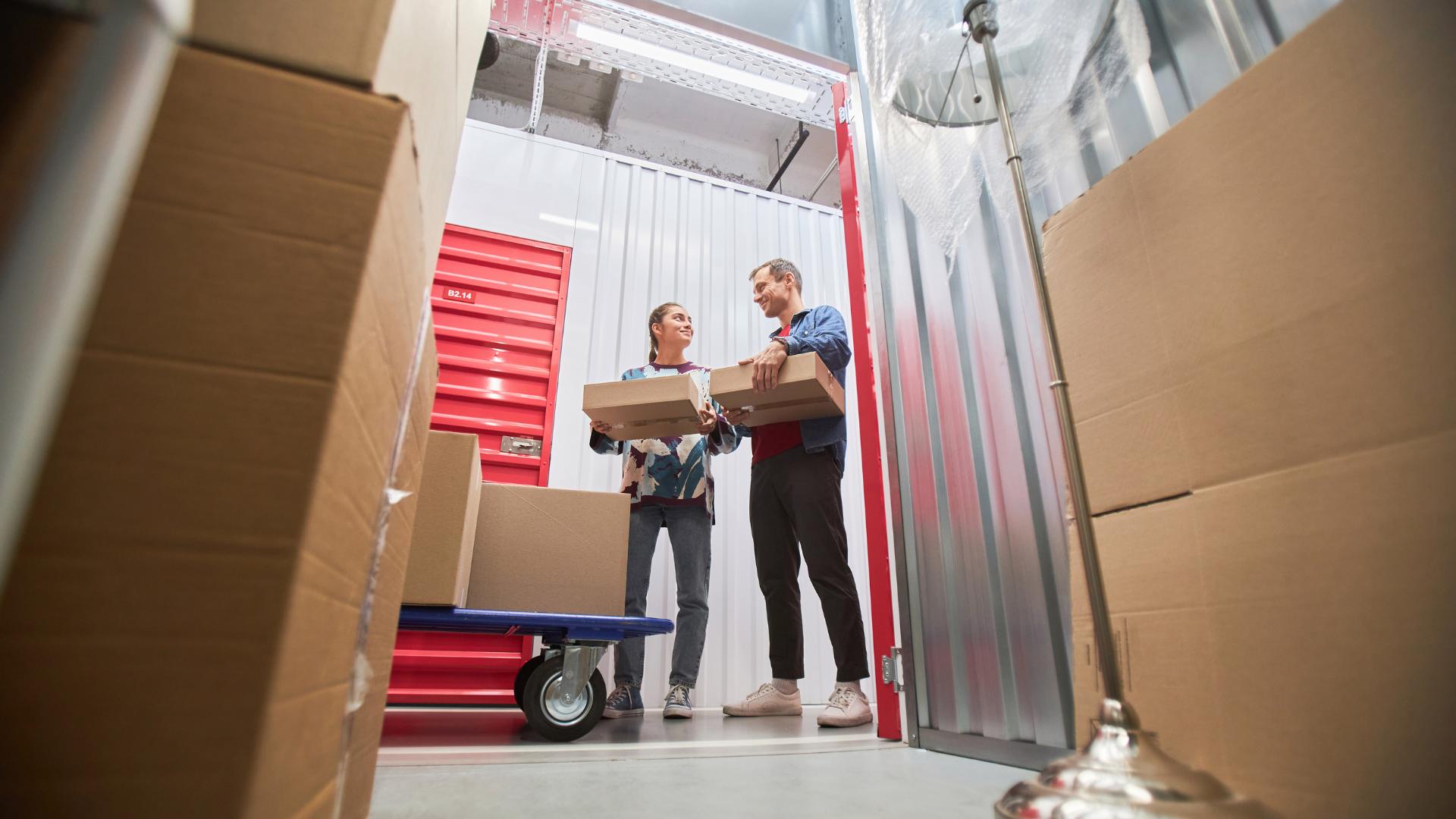 Two workers carrying boxes in a warehouse aisle with stacked red bins and large cardboard boxes