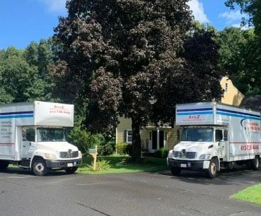 Two white moving trucks parked on a tree-lined street outside a house