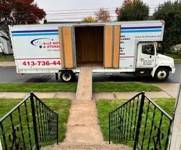 Moving truck with open cargo door and ramp parked in front of a house, seen from a front walkway.