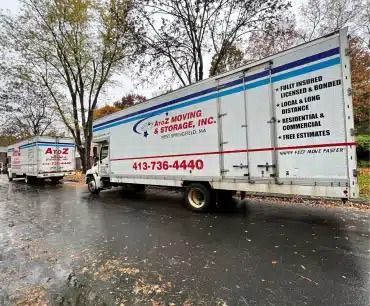 White moving truck on wet street, with another truck ahead in a rainy roadside setting.