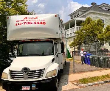 White moving truck parked by a house on a residential street, with blue trash bins and a chain-link fence.