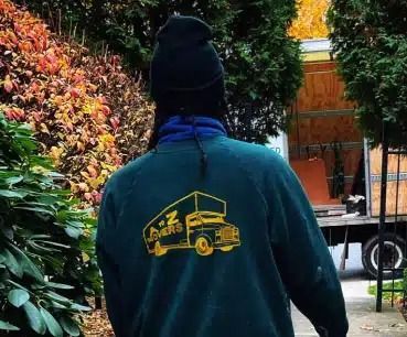 Person in black beanie and blue jacket with yellow truck graphic, seen from behind outdoors by foliage.
