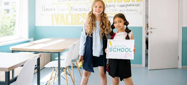 Two students standing in a bright classroom, holding a sign that says “Back to School.”
