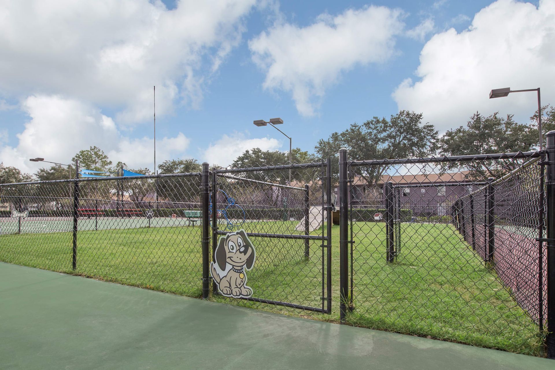 Fenced dog park with green grass, gate, and cutout dog. Blue sky, tennis court.