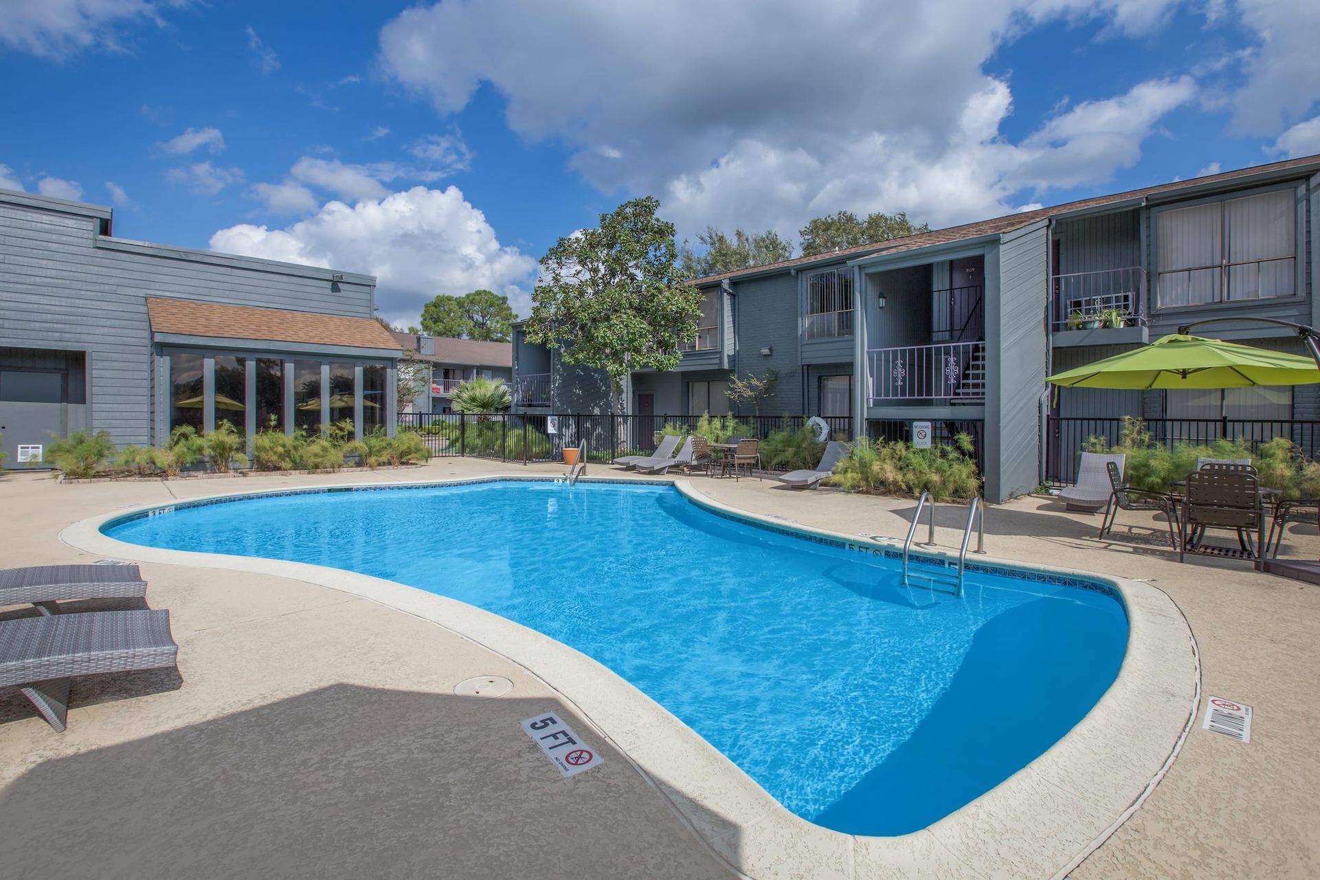 A swimming pool surrounded by a concrete patio, next to a two-story apartment building, under a blue sky.