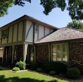 Two-story Tudor style home with beige stucco, dark brown beams, brick lower level, and shake shingle roof.