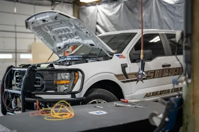 White sheriff's pickup truck with open hood in a garage. Tools and wires visible on a table.