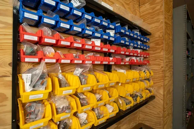 Wall-mounted storage bins in blue, red, and yellow, filled with parts, organized by color on a plywood wall.