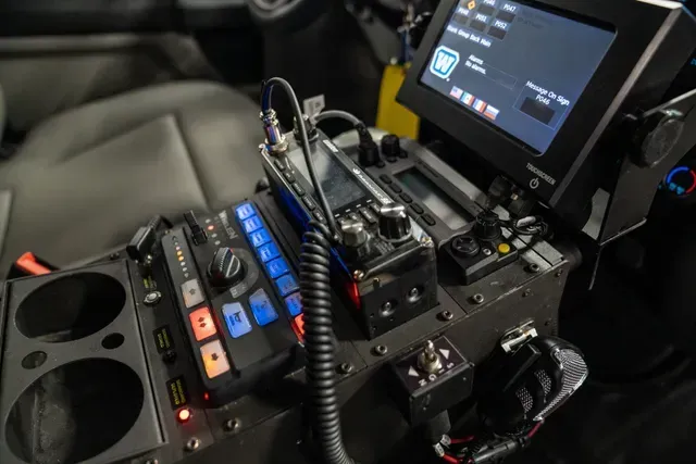 Dashboard of a police car with radio, screen, and control buttons, various colors, wires.