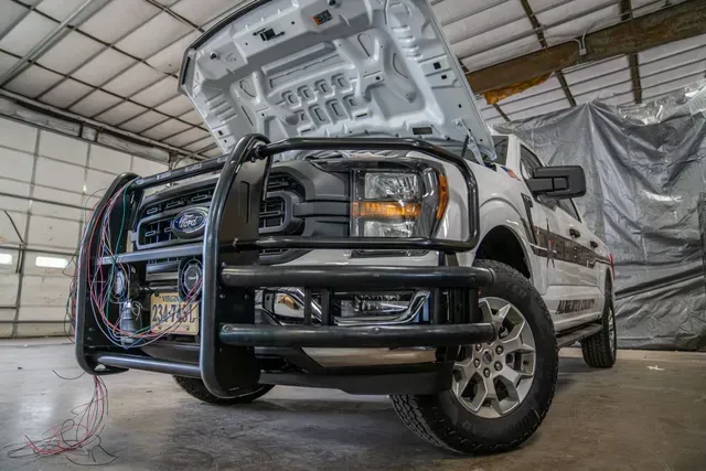 White pickup truck with hood up in a garage, a bull bar on the front, wires exposed.