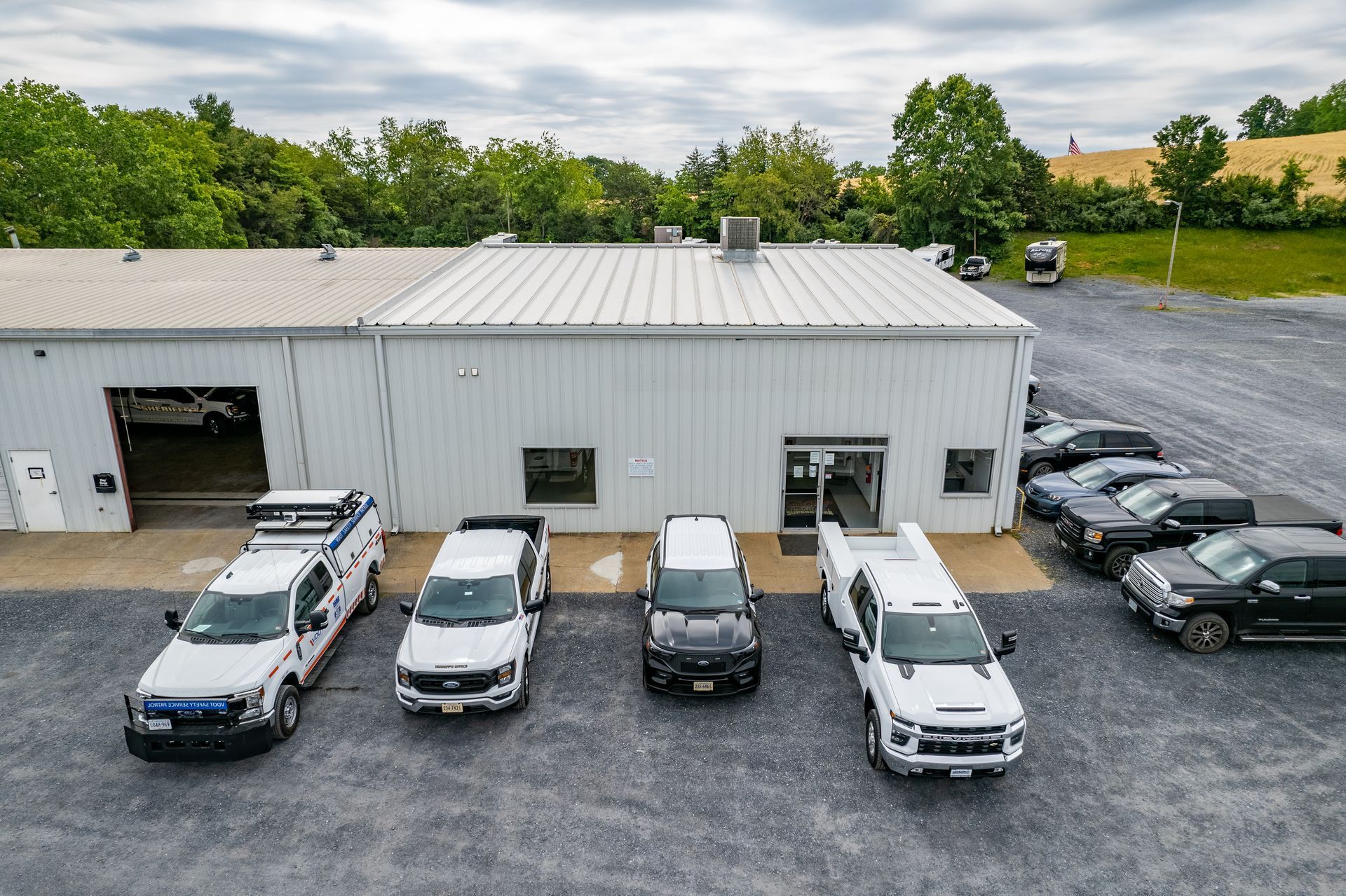 White building with several work trucks parked in front, gravel lot.
