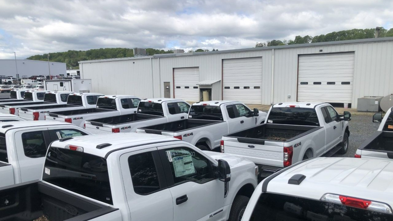 White pickup trucks parked in a lot in front of a white building with closed garage doors, under a cloudy sky.