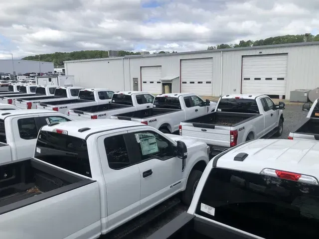 White pickup trucks parked in a lot in front of a white building with garage doors. Overcast sky.