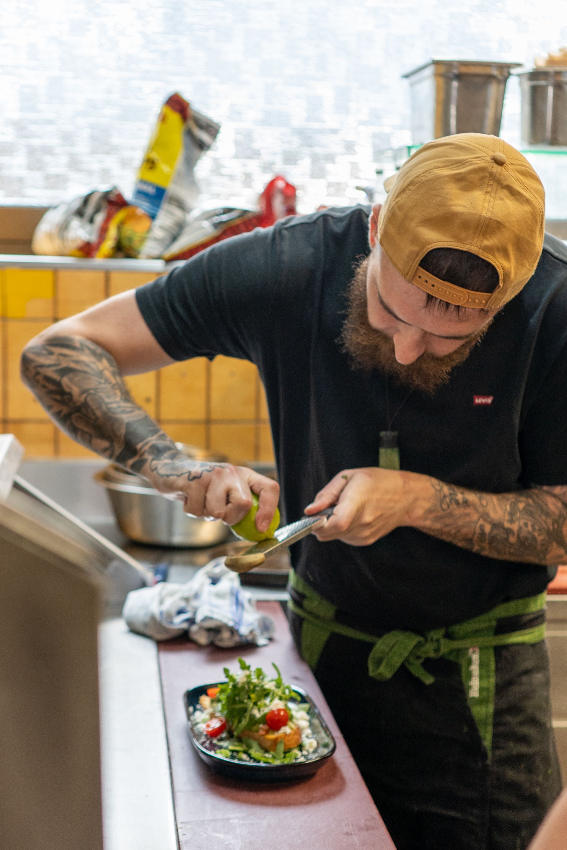 Un chef presse du citron vert sur une salade, portant une casquette et un tablier, dans une cuisine.