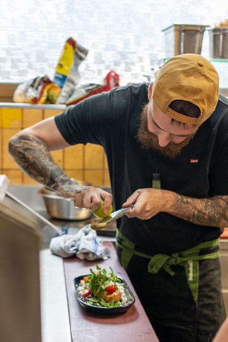 Un chef presse du citron vert sur une salade, portant une casquette et un tablier, dans une cuisine.