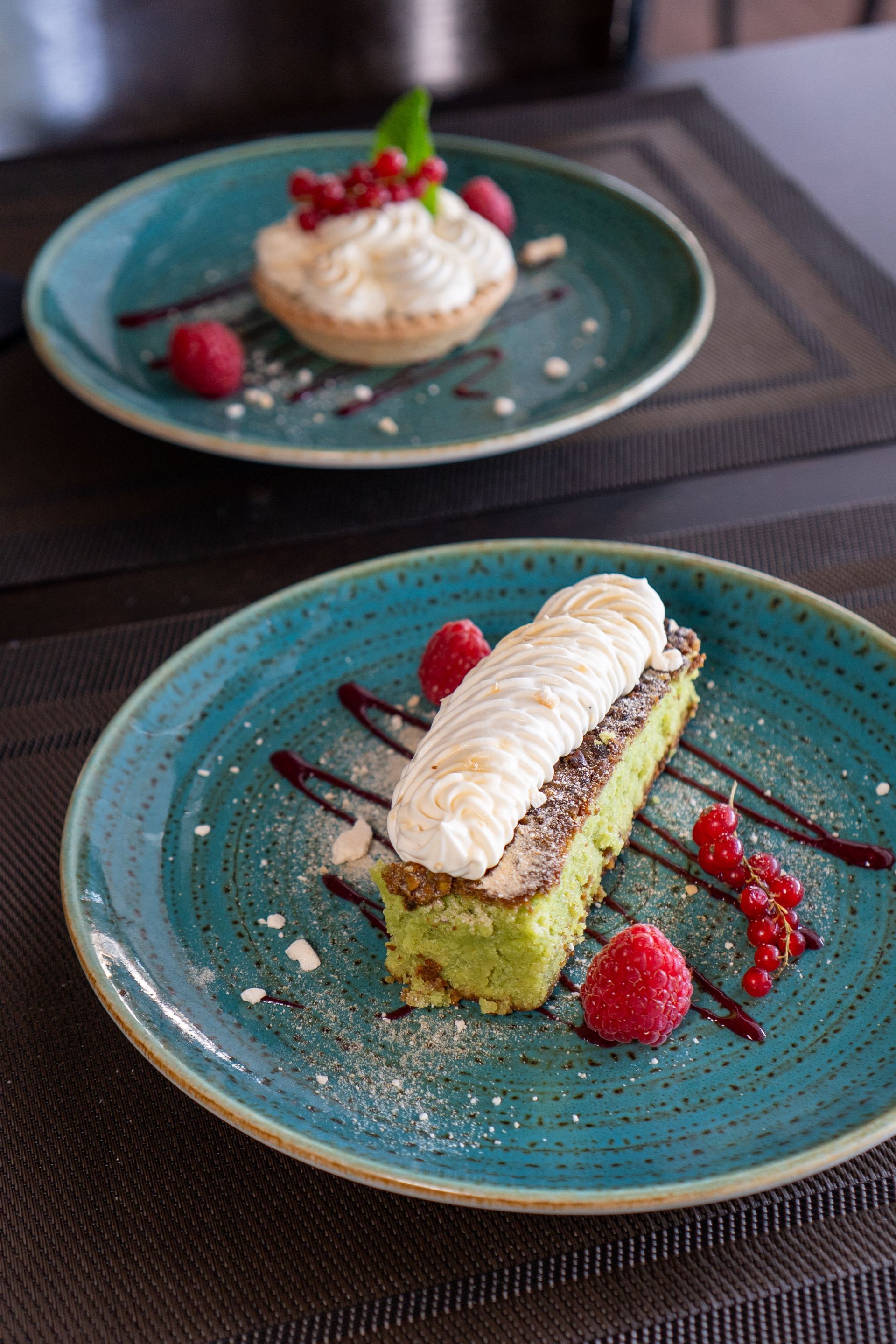 Deux assiettes avec desserts : gâteau vert et tarte garnis de crème, de fruits rouges et de sauce.
