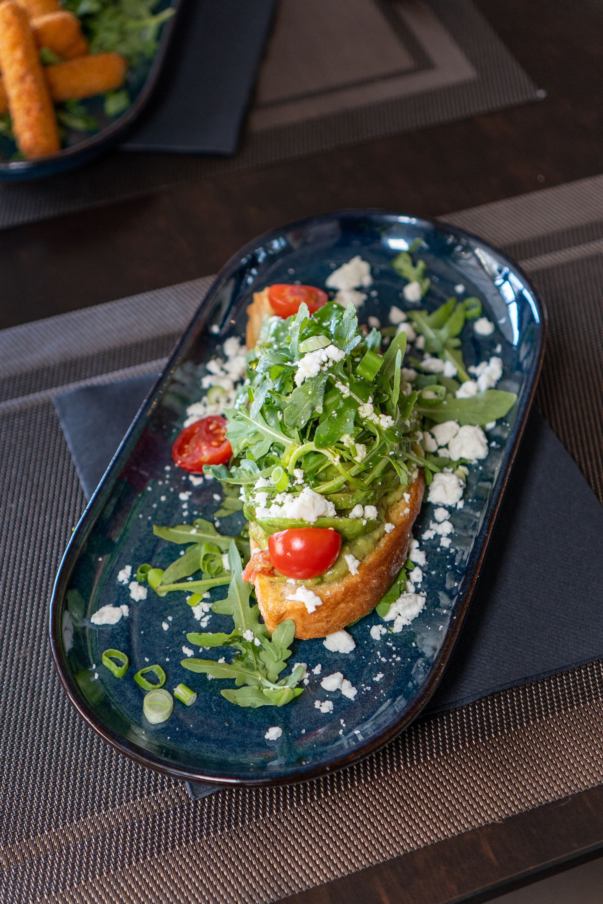 Tranche de pain grillée garnie d'avocat, de roquette, de tomates et de feta sur une assiette ovale bleue.