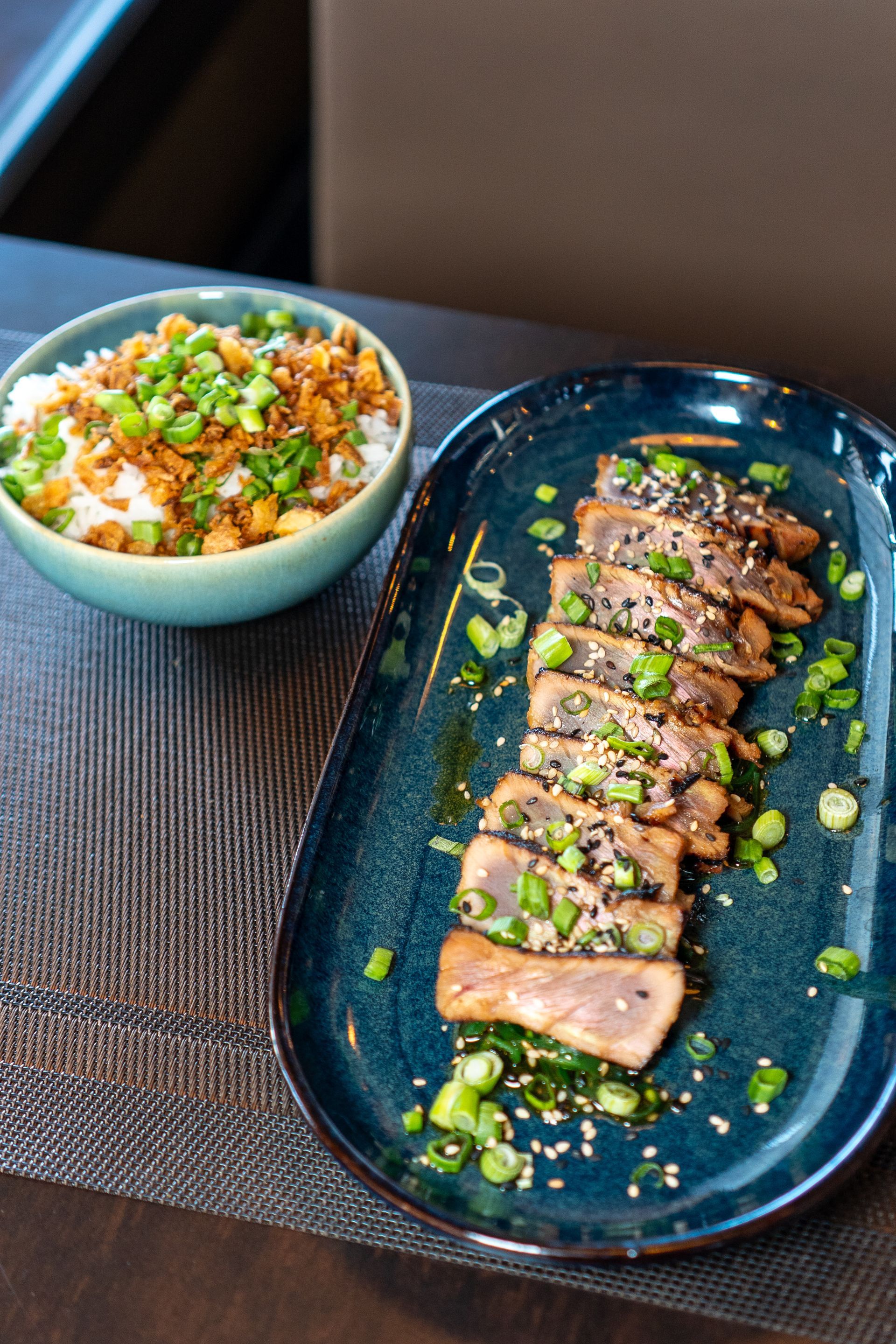 Tranches de thon poêlées sur un plateau bleu avec bol de riz, oignons verts, graines de sésame sur une table.