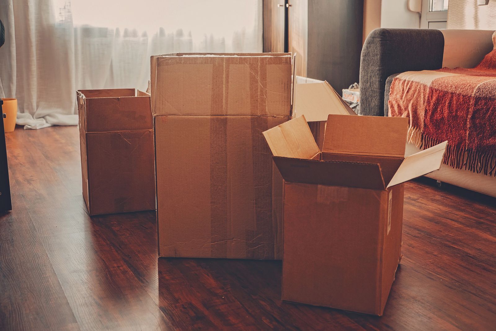Cardboard moving boxes on a hardwood floor in a room, with a couch and windows in the background.