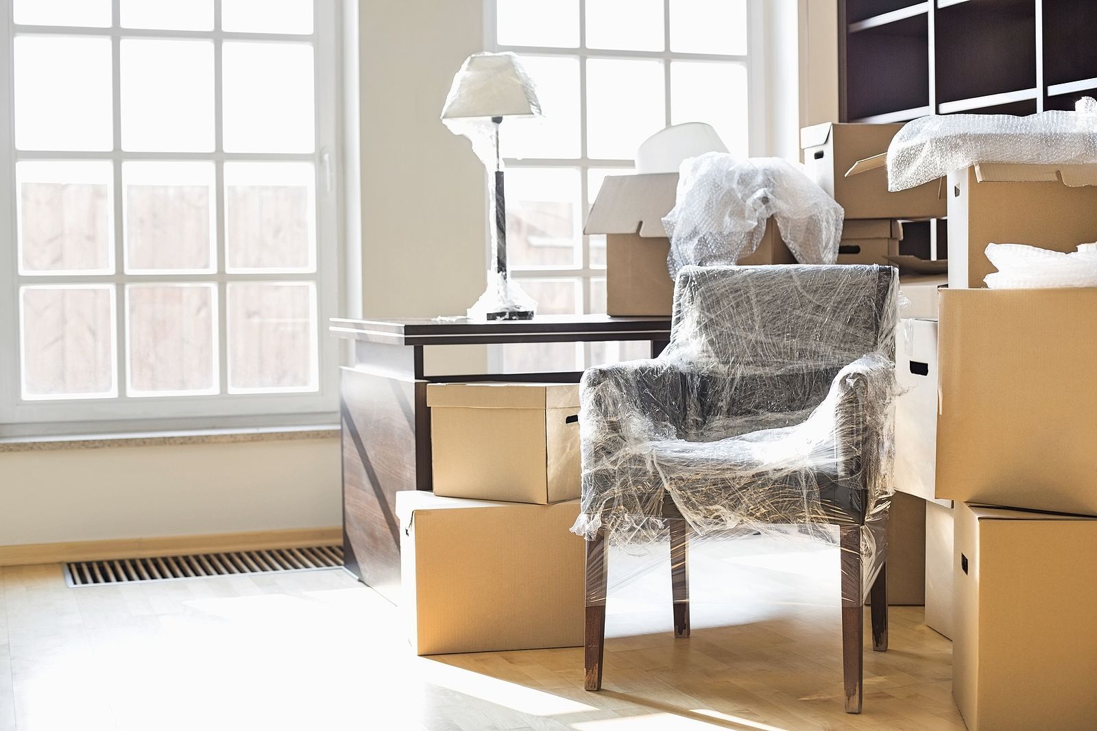 Room with packed boxes, wrapped furniture, and desk by windows, preparing for a move.