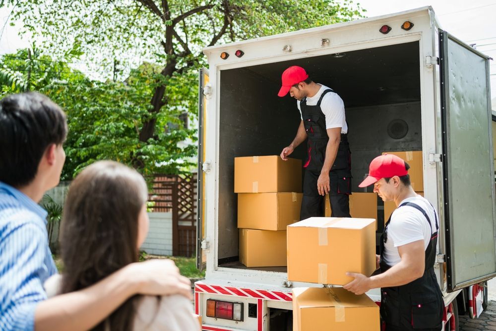 Two movers loading boxes into a truck as a couple watches; outdoors, sunny.