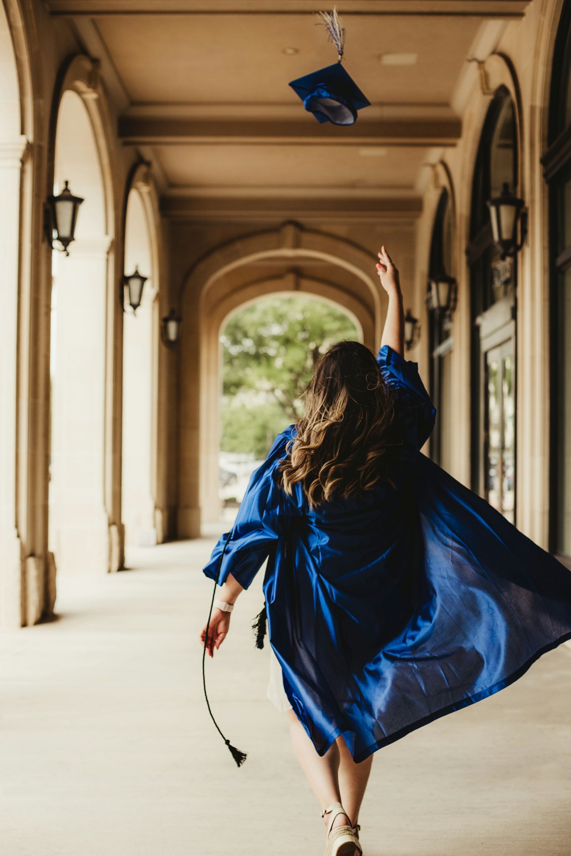 Person in blue graduation gown throws cap in air, walking through arched walkway.