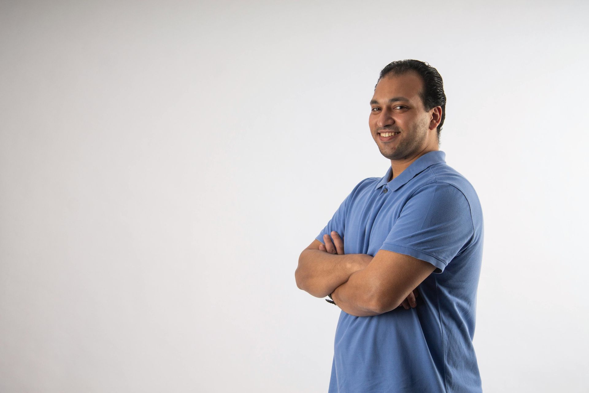 A man in a blue shirt is standing with his arms crossed and smiling.