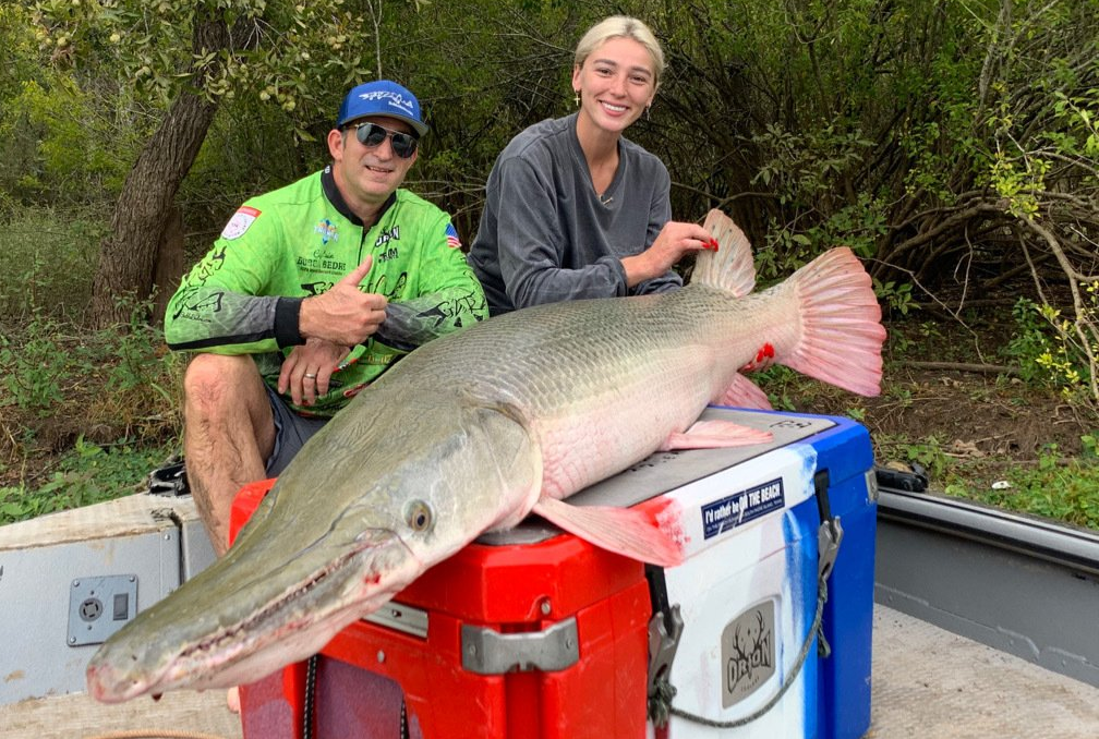 Two people pose with a large fish on a cooler. The fish is long with a pointed snout and pink fins.
