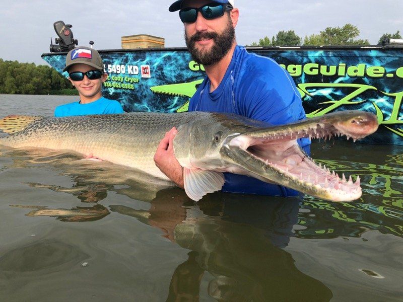 Man and boy holding a giant alligator gar in water, smiling, boat in background.