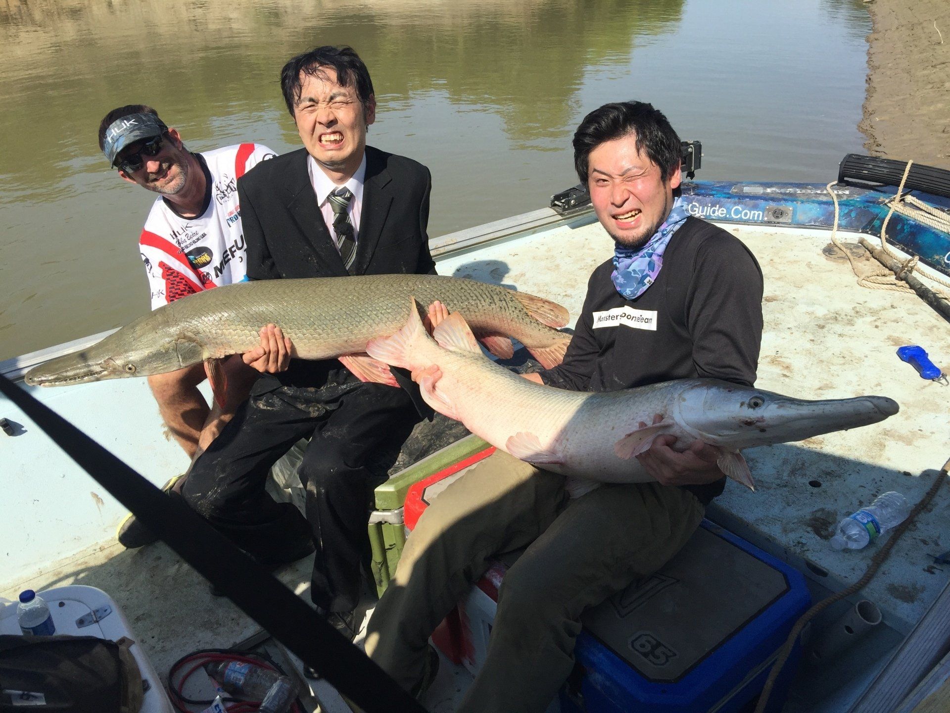 Three men on a boat hold large fish, smiling. River setting, sunny. One wears a suit.