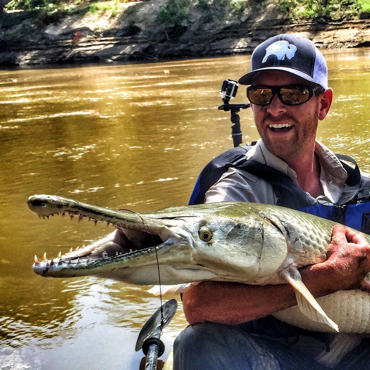 Man holding a large alligator gar fish, smiling, in a boat on a river.