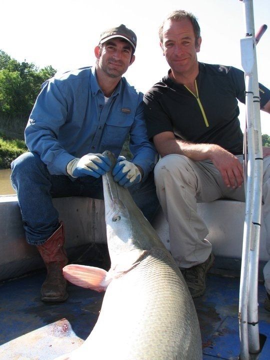 Two men on a boat holding a large fish. One man wears blue, the other black. Outdoors, daytime.